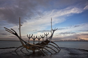 photo of the sun voyager, a sculpture in Reykjavik Iceland. Dark clouds are overhead and sunlit mountains are in the background.