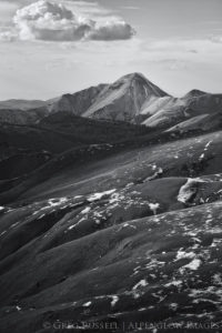 black and white photograph of a white mountain with a grassy hillsides in the foreground. Small patches of show are visible and clouds are in the sky.