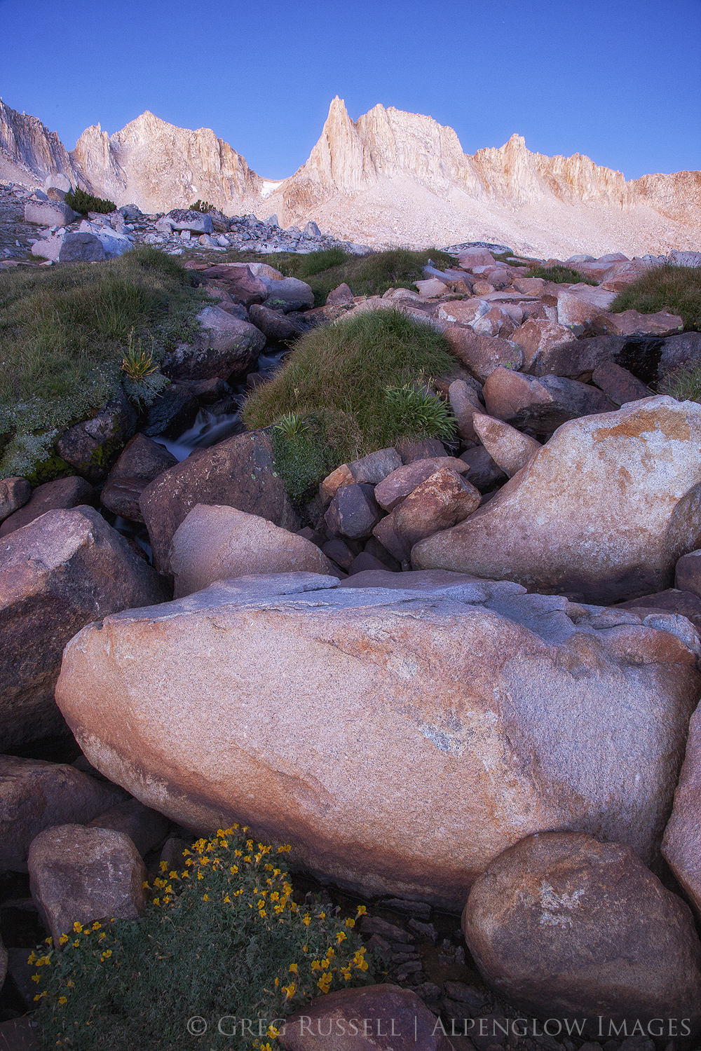 predawn light makes high Sierra Nevada peaks glow; a waterfall runs out from a lake beneath the peak.